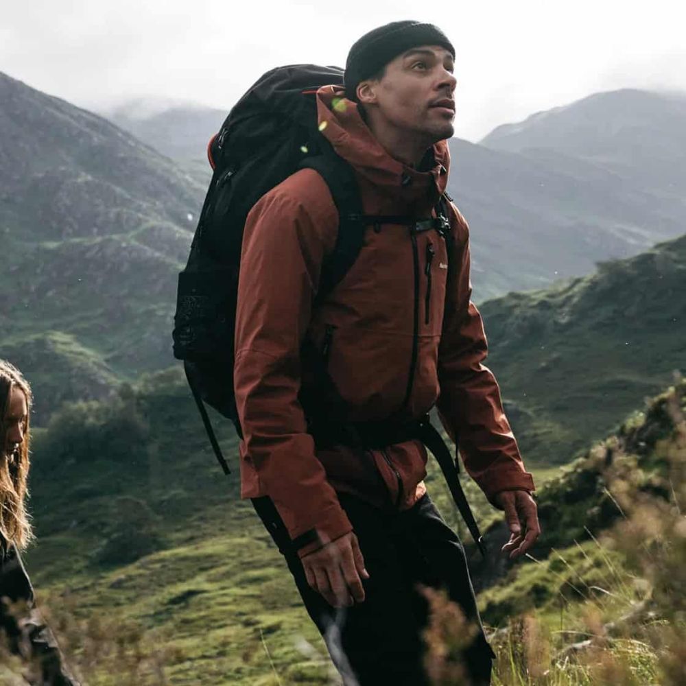 Person hiking in a mountainous landscape wearing a brown jacket and black backpack.